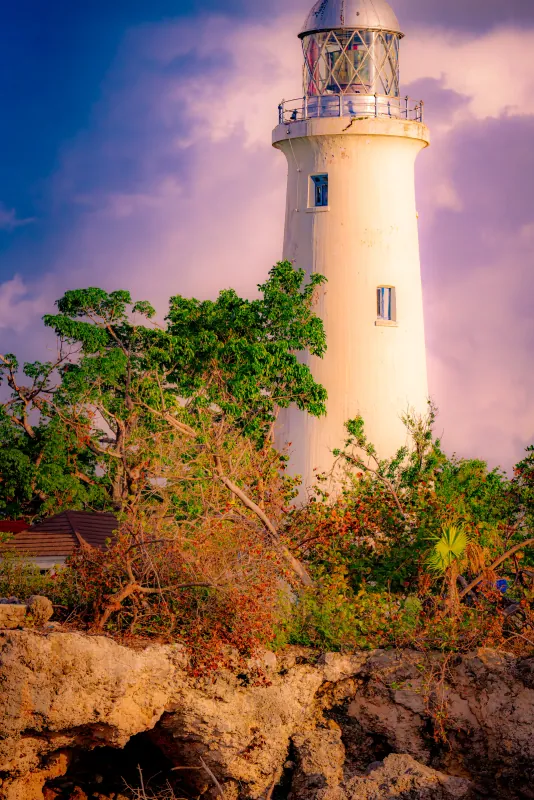 Negril Lighthouse in Jamaica glowing against a purple and pink dusk sky, tropical trees in the foreground