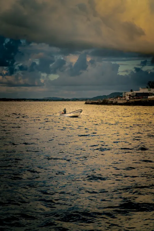 Small white boat on dark rippling water beneath heavy storm clouds along a tropical shoreline