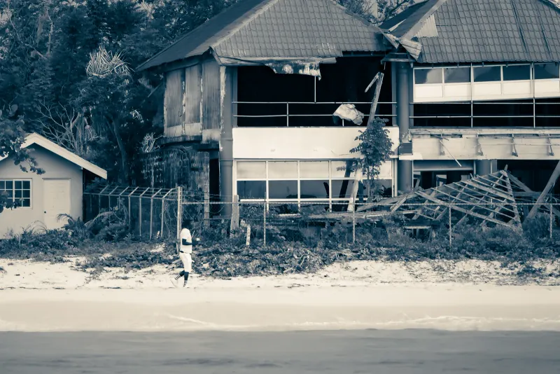 Black-and-white view of a storm-damaged beachfront building with a lone figure walking the sand
