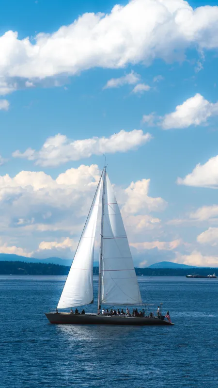 White-sailed sailboat heeling on blue water with mountains and cumulus clouds behind
